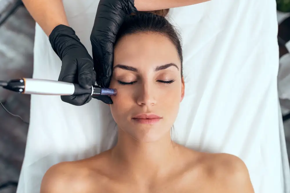 A provider performs microneedling on a woman’s cheek using a handheld device for Enhanced Wellness in Derby Kansas. The woman lies on a treatment bed with eyes closed while the provider wears black gloves.