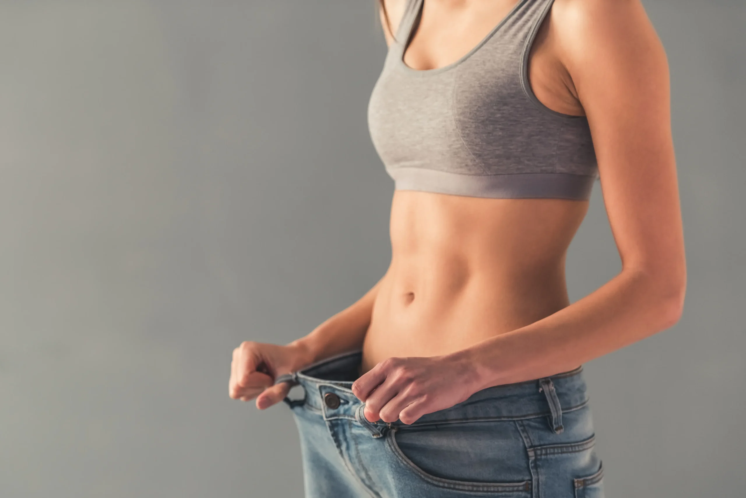 Woman showing loose jeans at the waist to highlight weight loss results for Enhanced Wellness in Derby Kansas. The image focuses on her midsection in a sports bra against a neutral background.
